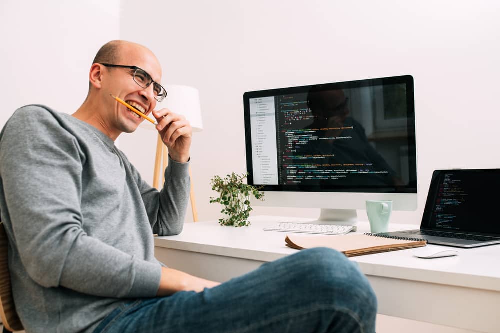 Programador sonriendo y mordiendo un lápiz frente a un monitor con código, representando el mito de la codificación en vivo en entrevistas.
